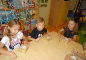 Czwórka dzieci siedzi przy stole na którym przed każdym dzieckiem leży kartonowe pudełeczko z nałożonym gumkami. Dzieci w ręku trzymają ołówki, którymi dotykają strun z gumek.